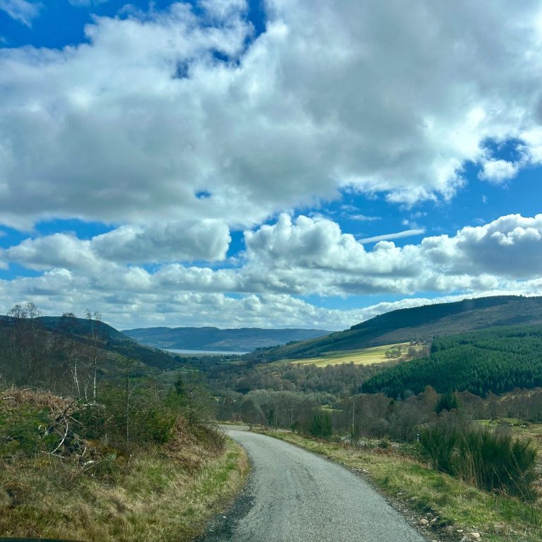 looking from the hills over Drumnadrochit with Loch Ness in the background.  The sky is blue and the clouds are white.