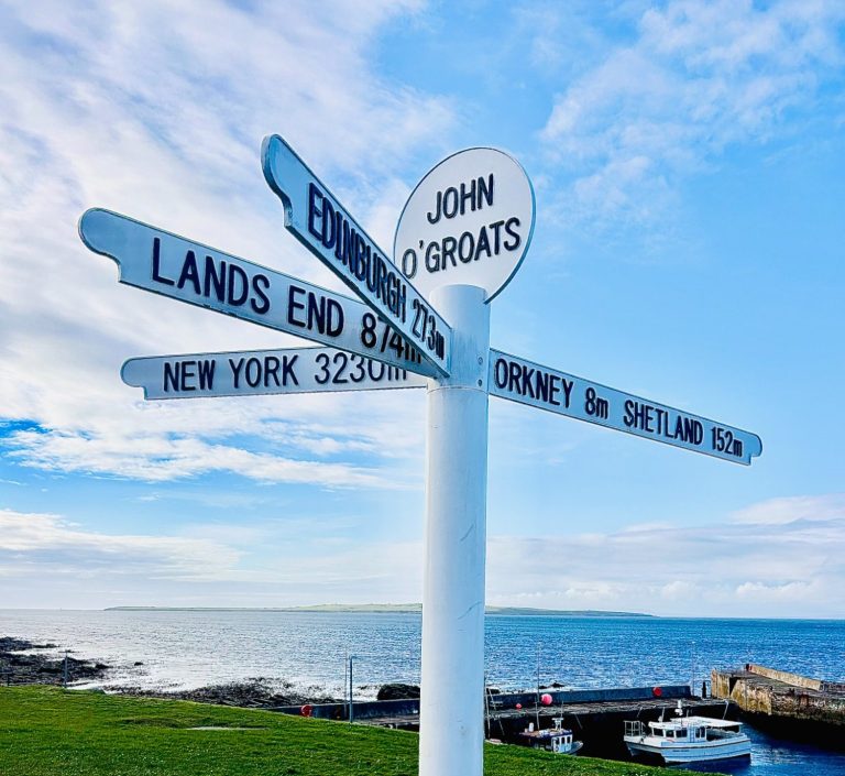 john O'Groats sign on NC500 with sea and harbour behind